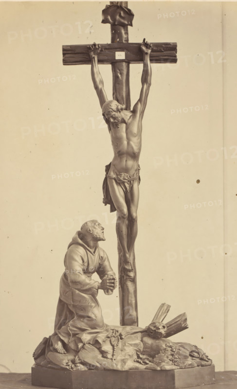 Palm wood crucifix from the collection of CJ Vervloet in Brussels, displayed at an exhibition on religious objects from the Middle Ages and Renaissance in 1864 in Mechelen, photograph, page, number: '9', photographer: Joseph Maes, Mechelen, 1864, paper, albumen print, height 251 mm x width 153 mm, altarcross, crucifix, piece of sculpture, reproduction of a piece of sculpture (wood, eg: panel, arts), Period, A monochrome photograph of a sculpture showing a crucified male figure on a wooden cross with his head bowed and a cloth around his waist, his arms outstretched and hands nailed to the crossbeam; below him a robed kneeling man looks upward with clasped hands, positioned on a rocky base strewn with sacks and a string of beads; a skull rests near broken wooden fragments at the base; the scene is framed against a plain background.