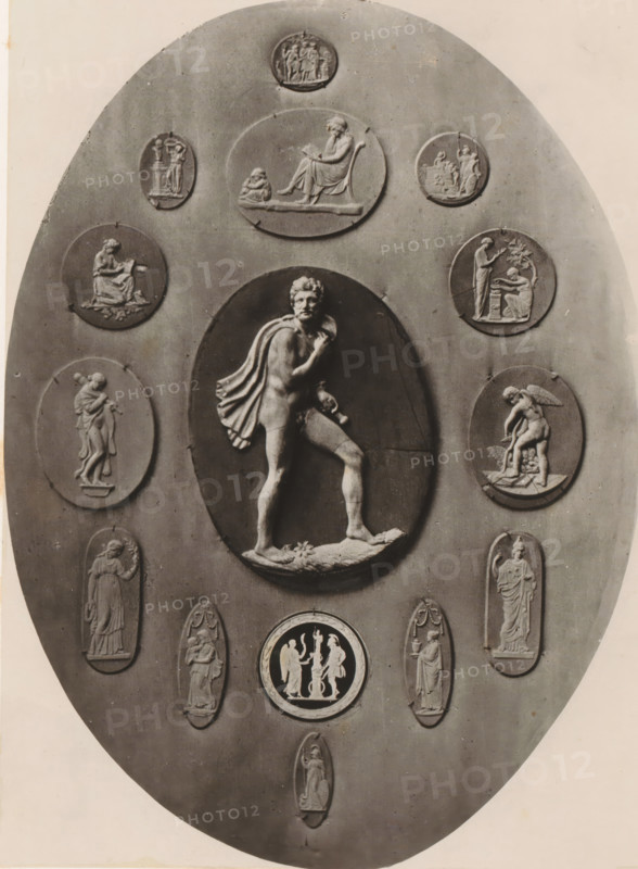 Medallion and fourteen cameos, photograph, page, photographer: Joseph Cundall, c. 1868 - in or before 1873, paper, carbon print, height 361 mm x width 270 mm, sculpture (relief, sculpture), carving, applied arts, An oval plaque with a central raised oval relief depicting a partially draped male figure standing with one foot on a rocky base and a cloak over his shoulder, surrounded by multiple smaller oval and circular relief medallions showing various classical-style figures in seated, standing, and interacting poses, all in a monochrome stone or plaster finish against a smooth background.