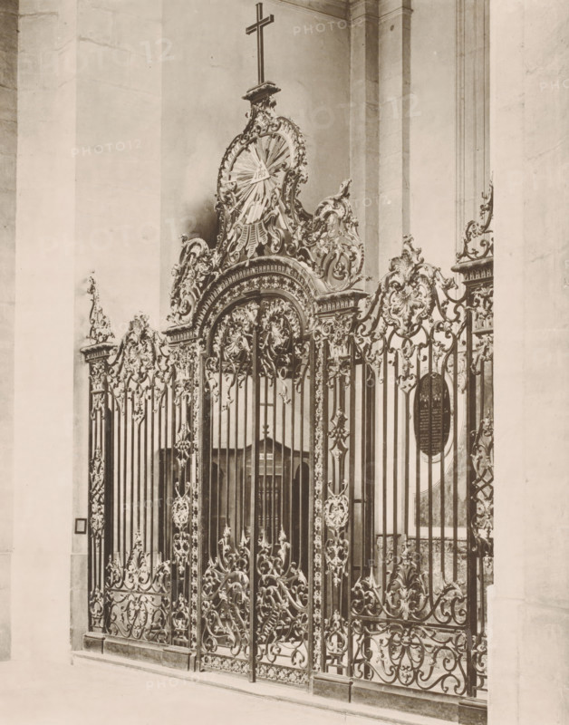 Chapel in the Notre-Dame de Saint-Sigisbert in Nancy, photomechanical print, This print is part of a cover with 103 prints on 101 pages and a title page., inscription: 'Cathédrale - primatiale, Chapelle dite des évêques, ', cliché maker: Royer & Cie., Nancy, publisher: Armand Guérinet, Paris, c. 1891 - in or before 1896, photographer, paper, collotype, photoprint, height 401 mm x width 298 mm, parts of church interior: chapel, A tall ornate wrought iron gate with intricate scrollwork and floral motifs fills the foreground, topped by a decorative arch and cresting elements that include a radiating sunburst design and a cross above it; behind the gate a stone interior with high smooth pillars and a recessed doorway is visible, and the wrought metalwork continues along the sides with repeating vertical bars and ornamental panels, all shown in a monochrome photograph with soft lighting and subtle shadows.