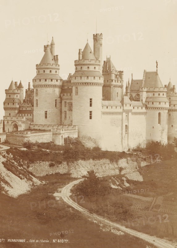 Vue d'Ensemble - Côté de l'entrée, View of the castle of Pierrefonds, France, Chateau de Pierrefonds, photograph, page, number: 'PL. 1', inscription: 'No. 410bis PIERRE FUND. - Côté de l'Entrée. No. 410bis.', photographer: Séraphin-Médéric Mieusement, Pierrefonds, publisher: Ducher & Cie., c. 1870 - in or before 1875, Séraphin-Médéric Mieusement, Ducher & Cie., photographer, publisher, paper, albumen print, albumendruk, height 351 mm x width 250 mm, castle, Pierrefonds Castle, A large stone castle with multiple rounded towers and conical roofs rises on a cliff, showing crenelated battlements and narrow vertical windows; ornate rooflines and sculptural details appear on some towers, and connecting walls feature small rectangular openings and decorative stonework; a winding dirt path leads up the slope toward the castle, bordered by grassy and rocky terrain with patches of low vegetation; the foreground shows layered earth and stone forming the cliff face beneath the castle.