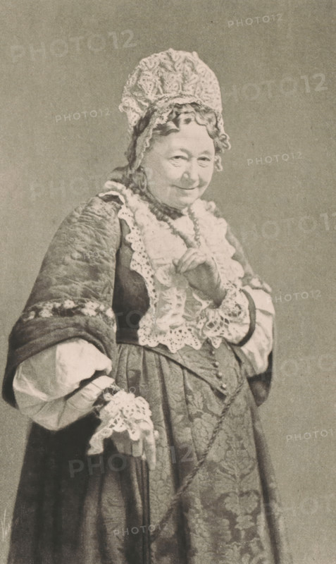 Mrs Stirling, Portrait of Mary Anne Stirling in the role of a sister, page, photomechanical print, inscription: '(THE NURSE.)', photographer: Henry van der Weyde, cliché maker: The Art Reproduction Company (possibly), publisher: Virtue & Co., c. 1885 - in or before c. 1890, paper, photoaquatint, height 202 mm x width 128 mm, Print on third unnumbered page after cover page., Person, Mary Anne Stirling, historical persons - BB - woman, portrait of actor, actress, An older woman wearing a tall, lace-trimmed cap and a dress with ornate lace ruffles and patterned fabric is shown from head to mid-thigh, her hair visible under the cap and her face showing a slight smile, one hand raised near her chest with fingers gently curved and the other resting lower with a lace cuff draped over it, the clothing featuring layered sleeves, textured shawl, and a beaded necklace against a plain background.