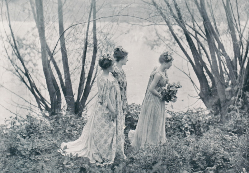 Landscape with three women with flowers in their hair, page, photomechanical print, photographer: Constant Puyo, cliché maker: Jean Malvaux, Brussels, c. 1895 - in or before 1900, paper, autotype, height 154 mm x width 215 mm, flowers, landscapes, adult woman (three persons), Three women in long flowing dresses stand in a natural setting among tall bare tree trunks and low foliage, viewed from the side with their profiles visible; two stand close together while the third is slightly ahead holding a bouquet of flowers, each with hair styled up and small flower adornments, and the scene has a soft, muted monochrome tone.