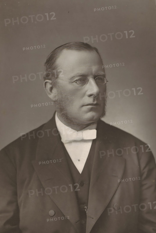Portrait of Edmond Warre, The Rev. Dr. Warre, page, photomechanical print, inscription: '57 & 61, Elbury Street, London.', photographer: Downey, William & Daniel, London, cliché maker: anonymous, c. 1880 - in or before 1890, paper, Woodburytype, height 141 mm x width 95 mm, Person, Edmond Warre, university organization, A sepia-toned portrait of a man shown from the chest up wearing a dark suit jacket over a dark vest and a light-colored shirt with a light bow tie; he has short, neatly combed hair, full sideburns, and small round eyeglasses, and his expression is neutral while he looks slightly to the side against a plain background.