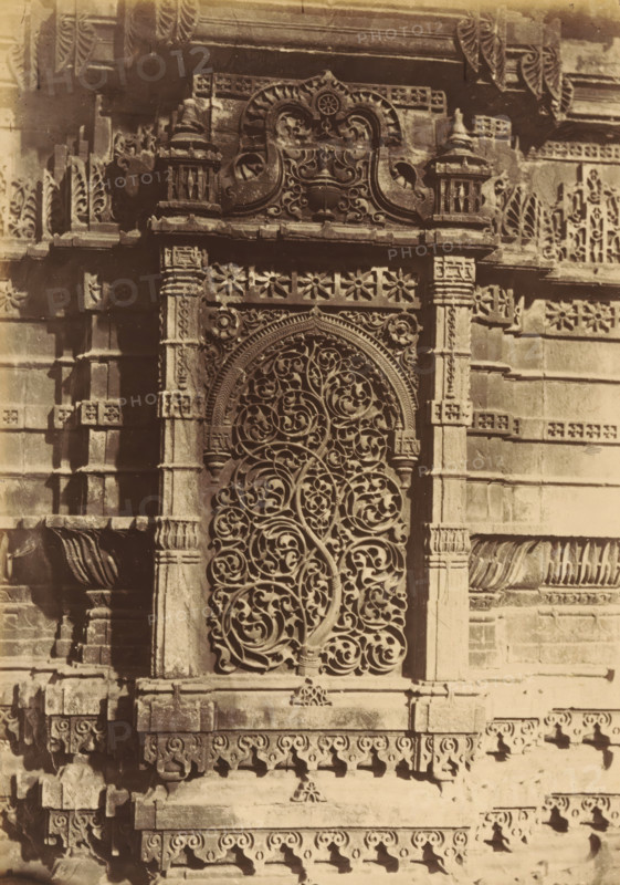 The Shápoor Mosque.- Niche from the base of the Northern Minaret, Decorations on a base of a minaret of the Shapoor Mosque in Ahmedabad, depicting a tree, photograph, page, number: '107.', photographer: Thomas Biggs, Ahmedabad (city), c. 1856 - in or before 1866, Gujarat, paper, albumen print, height 186 mm x width 136 mm, ornament derived from plant forms, temple, shrine, Islam, Mohammedanism, base (, column, pillar), A close view of an ornate stone wall section featuring a tall arched window with an intricate carved grille of intertwining vine and leaf patterns, flanked by decorated stone pilasters and framed by a carved arch with floral and scroll motifs; above the arch is a carved panel with symmetrical scrollwork and ornamental finials, and the surrounding masonry shows layered cornices, patterned friezes, and repeated floral and geometric reliefs with areas of shadow and weathering.