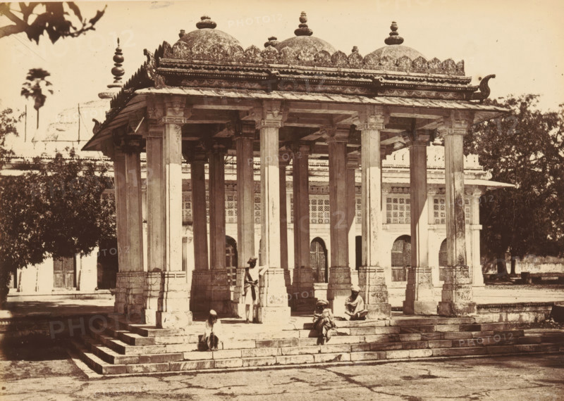 Sirkhej.- The Pavilion, and the Tomb of Gunj Buksh, Pavilion and the tomb of Ahmed Khattu Ganj Baksh at Sarkhej Roza near Ahmedabad, photograph, page, number: '43.', photographer: Thomas Biggs, Sarkhej Roza, c. 1856 - in or before 1866, Gujarat, paper, albumen print, height 136 mm x width 187 mm, grave, tomb, A stone pavilion with a raised platform and a colonnade of tall square pillars supports an ornate flat roof topped by multiple domes and decorative finials, with carved cornices and scalloped edging along the roofline; steps lead up to the platform and a few seated and standing figures in traditional clothing are on and near the steps; behind the pavilion a building with arched openings and patterned window screens is visible, and trees frame parts of the scene.