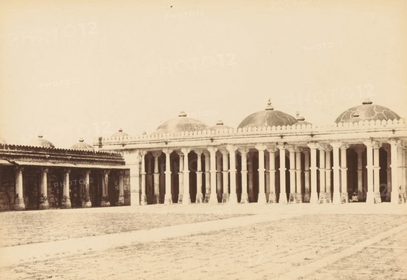 View of a mosque at Sarkhej Roza near Ahmedabad, Sirkhej.- The Mosque, fro the North-east, photograph, page, number: '48.', photographer: Thomas Biggs, Sarkhej Roza, c. 1856 - in or before 1866, Gujarat, paper, albumen print, height 136 mm x width 187 mm, temple, shrine, Islam, Mohammedanism, A long colonnaded building with a series of tall, evenly spaced pillars supporting a horizontal entablature and a row of domed roofs above, the domes topped with small finials and showing visible surface patterns, an open arcade beneath the entablature with shadowed recesses and visible architectural details on the pillars and arches, a broad open ground in front with faint textured paths across it, and a person seated near one end of the colonnade.