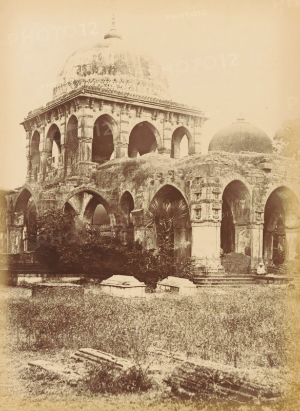 Butwa, - The Tomb of Kootub-i-Alum, Tomb of Kootub-i-Alum in Batwa near Ahmedabad, photograph, page, number: '60.', photographer: Thomas Biggs, Ahmedabad (district), c. 1856 - in or before 1866, Gujarat, paper, albumen print, height 202 mm x width 148 mm, Person, Kootub-i-Alum, grave, tomb, A sepia-toned photograph of an old stone building with large rounded domes and multiple arched openings, showing weathered and textured stonework, carved details around the arches, and vegetation growing on and around the structure; a foreground of grassy ground with scattered stone slabs and low tomb-like structures sits before the building, and a single person wearing light clothing and a hat sits on steps near one of the arches.