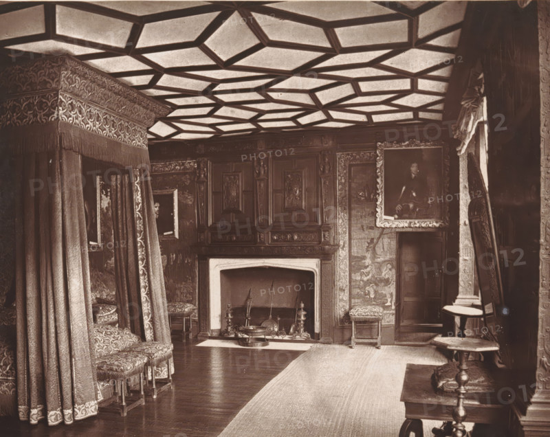 Interior of a bedroom at Knole House, The spangled bed-room, Knole, page, photomechanical print, photographer: Stephen Thompson, Knole House, c. 1866 - in or before 1876, printmaker, Kent, paper, Woodburytype, height 147 mm x width 185 mm, bedroom, interior of the house, A richly furnished interior with a patterned coffered ceiling, ornate draperies forming a canopy over a bed area at left, upholstered stools with decorative fabric and fringe, a carved wooden fireplace surround with a mantel, and a hearth containing fireplace tools andirons and a low decorative brazier; carved wooden paneling and framed paintings hang on the walls, one large portrait in an ornate frame at right, a tapestry or wall covering with figures beside the fireplace, a narrow rug running along a polished wooden floor, a small round table with a turned pedestal and an upholstered footstool nearby, and heavy curtains and a carved doorway visible at the far right.