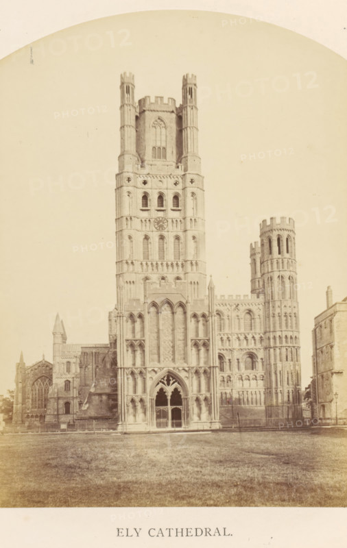 Ely Cathedral, photograph, page, Ely, c. 1858 - in or before 1869, photographer, Cambridgeshire, paper, albumen print, height 183 mm x width 127 mm, Photo on frontispiece., church (exterior), facade (of house or building), A large stone cathedral with a central tower flanked by a round tower and lower aisles stands on a grassy foreground; the central facade shows tall arched windows and decorative stonework with a recessed entrance featuring pointed arches and tracery, the main tower rising with multiple levels of narrow windows and a circular opening, the round tower to the side displaying vertical shafts and battlemented top, adjoining lower sections and buttresses extend to the left and right, and a paved area and fence run along the base of the building.