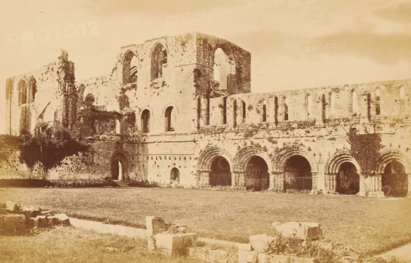View of the ruins of Furness Abbey, Furness Abbey, from the south-west, photograph, page, photographer: Garnett & Sproat, c. 1857 - c. 1867, Cumbria, paper, albumen print, height 106 mm x width 166 mm, ruin of church, monastery, A sepia-toned photograph of a large stone ruin with tall arched openings and a series of smaller arched arcades along its façade, showing weathered masonry and patches of climbing vegetation on the walls, with a grassy foreground scattered with stone blocks and remnants, and parts of the structure featuring large window openings and recessed doorways.