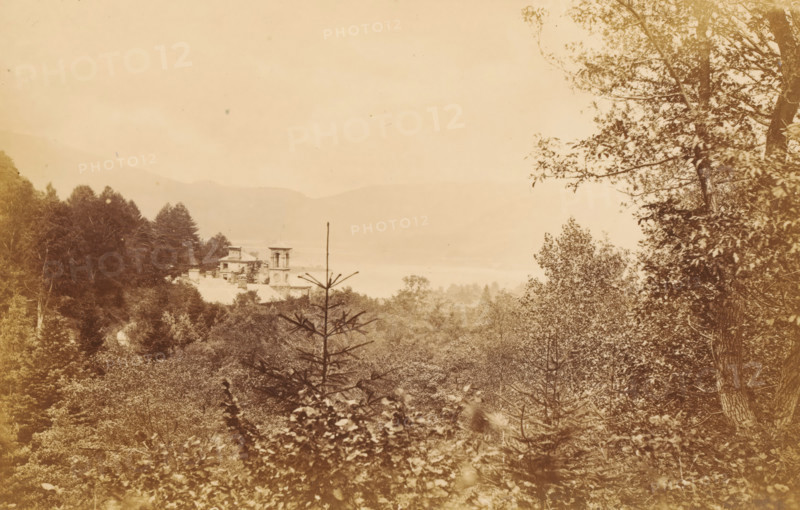 Ullswater, from Grisedale, View of Ullswater in the Lake District, photograph, page, photographer: Garnett & Sproat, Lake District, c. 1857 - c. 1867, paper, albumen print, height 105 mm x width 165 mm, lake, forest, wood, mountains, A sepia-toned landscape showing dense trees and foliage in the foreground and middle ground with a narrow conifer protruding centrally, a multi-tiered building with a square tower partially visible among the trees to the left of center, a calm body of water behind the building, distant hazy hills across the horizon, and leafy branches framing the right edge of the image.