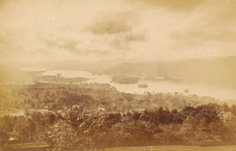 View over Windermere, Winander Mere, from Elleray, looking south, photograph, page, photographer: Garnett & Sproat, Lake District, c. 1857 - c. 1867, paper, albumen print, height 106 mm x width 163 mm, lake, Windermere, A wide sepia-toned landscape showing a lake with several small islands and peninsulas surrounded by rolling hills and tree-covered terrain, a foreground of dense trees and foliage, scattered buildings partially visible among the trees, a calm water surface reflecting the sky, distant layered hills fading into the background, and a cloudy sky with soft, diffuse light.