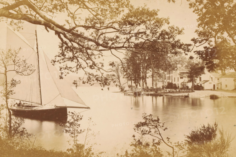 Sailboat on Lake Windermere near the ferry landing, Yachting at the Ferry, Winander Mere, photograph, page, photographer: Garnett & Sproat, Windermere, c. 1857 - c. 1867, Lake District, paper, albumen print, height 102 mm x width 154 mm, cottage, sailing-ship, sailing-boat, A calm waterfront scene with a sailboat moored near the left foreground, its tall mast and triangular sails prominent, and a person seated in its stern; large overhanging tree branches and leafy foliage frame the top and sides of the image, casting reflections on the water; a small wooden dock extends into the water toward the center right, with several rowboats tied nearby; a group of trees stands on a narrow point of land behind the dock; a cluster of multi-story buildings with pitched roofs and chimneys sits near the shore to the right, partially obscured by trees and shrubs; smooth water fills the central area, showing soft reflections of the boats, trees, and shoreline.