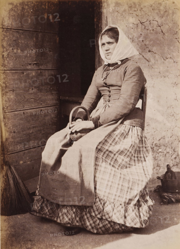 Portrait of an unknown woman on a chair, photograph, page, Italy, c. 1850 - in or before 1860, photographer, paper, albumen print, height 135 mm x width 96 mm, adult woman, historical persons not known by name - BB - woman, A seated woman wearing a headscarf and layered dress sits near a wooden door and a cracked plaster wall, holding a small rounded basket in her lap with both hands, her gaze directed slightly away from the camera, with a broom leaning against the door and a metal pot on the ground beside her.