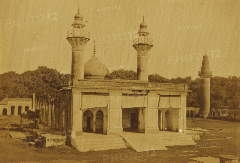 View of the Karbala Talkatora in Lucknow, Kurballa Talkatora, photograph, page, number: '50', photographer: Darogha Ubbas Alli, Lucknow, c. 1864 - in or before 1874, Oudh, paper, albumen print, height 105 mm x width 154 mm, temple, shrine, Islam, Mohammedanism, A sepia-toned photograph of a building complex with architectural elements including a domed roof and several cylindrical towers topped with ornate finials and balcony-like platforms; the foreground structure has a colonnaded façade with arched openings and a short flight of steps leading up to it, and portions of the walls show weathering and dark streaks; behind and beside the main building are dense clusters of trees and additional arched structures extending along one side; the ground in front shows patches of grass, stone slabs, and low rectangular stone blocks scattered around.