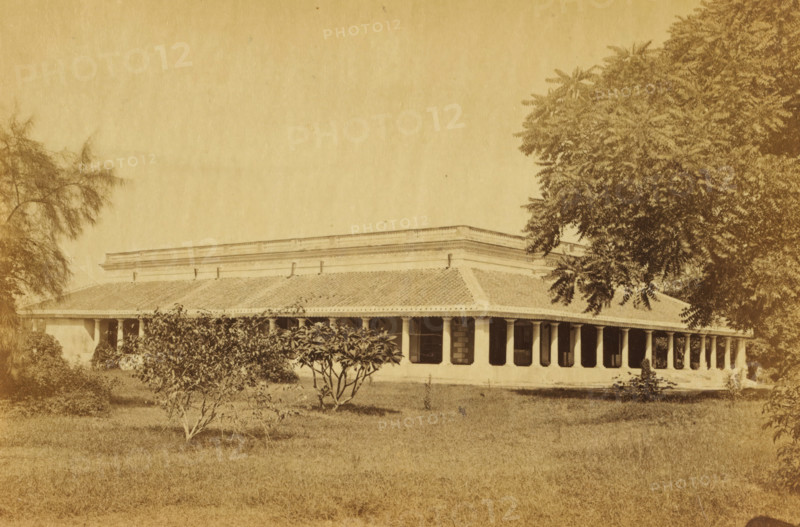 Darus-shafa, View of a hospital, Dar-us Shafa, in Lucknow, photograph, page, number: '8', photographer: Darogha Ubbas Alli, Lucknow, c. 1864 - in or before 1874, Oudh, paper, albumen print, height 105 mm x width 155 mm, hospital, A sepia-toned photograph shows a single-story building with a long tiled roof and a covered veranda supported by columns, dark window openings along the facade, and decorative cornice detail along the roofline; the building is set behind a grassy foreground with small leafy shrubs and trees, and larger trees frame the scene on both sides.