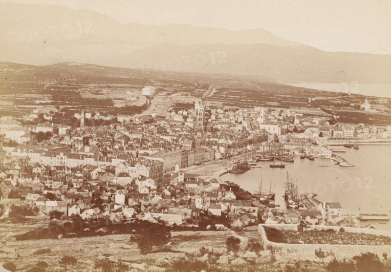 Spalato, View of Split, Spalato and seine Alterthümer, photograph, page, photographer: Franz Thiard de Laforest, Splitsko-Dalmatinska Županija, c. 1868 - in or before 1878, Yugoslavia, paper, albumen print, height 103 mm x width 148 mm, city-view in general, 'veduta', Split, A sepia-toned panoramic view shows a coastal town with densely packed buildings and narrow streets extending toward a waterfront where several sailing ships are moored along curved quays and piers; among the buildings are taller structures with towers and steeples rising above lower rooftops, and a long row of multi-story facades faces the harbor; beyond the town there are terraced fields and low stone walls stretching across a gently rolling landscape that leads to distant layered hills and mountains under a pale sky.