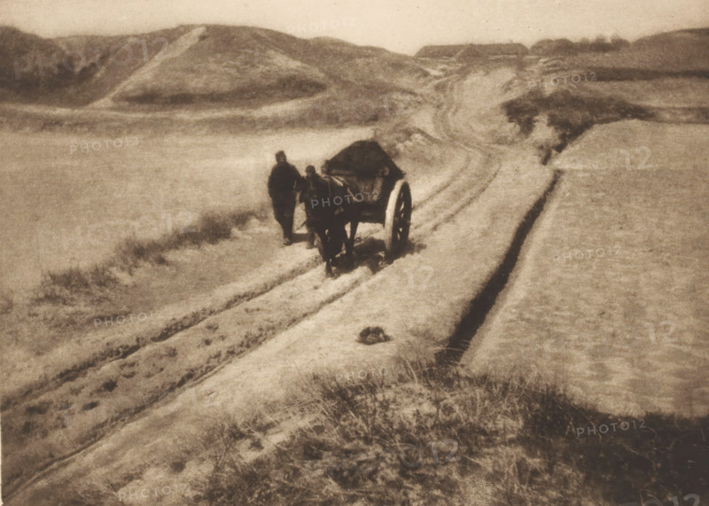 View of two unknown men with a cart on a path through the dunes, The road through the dunes, page, photomechanical print, photographer: James Craig Annan, cliché maker: Meisenbach, Riffarth & Cie., Berlin, publisher: Wilhelm Knapp Verlag, Halle an der Saale, c. 1897 - in or before 1902, Saxony-Anhalt, paper, photoaquatint, height 146 mm x width 203 mm, dunes (sea not visible), in the dunes, road, path, hand-cart, A grainy monochrome scene shows a sandy landscape with undulating dunes and a winding track cutting through the sand. Near the center a covered cart with large wheels stands on the track, accompanied by two upright figures close by. Tire or wheel ruts run along the path leading toward distant low hills. Sparse tufts of grass and scattered vegetation appear in the foreground and along the edges of the track. The overall tones are muted and sepia-like, with soft contrasts and a hazy sky above the dunes.