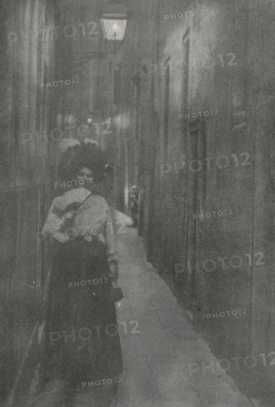 Gasse in Venedig, Portrait of an unknown woman with fan and hat in an alley in Venice, page, photomechanical print, photographer: Alfred Kirstein, Venice, cliché maker: Meisenbach, Riffarth & Cie. (possibly), Berlin, publisher: Wilhelm Knapp Verlag, Halle an der Saale, c. 1897 - in or before 1902, Saxony-Anhalt, paper, autotypie, height 212 mm x width 145 mm, Print seven pages after page 102., adult woman, anonymous historical person depicted, A narrow, dimly lit alley with tall walls on either side and a light fixture mounted high above; a woman stands in the foreground wearing a dark skirt and a light blouse, holding a folded hand fan partly in front of her face and carrying a small handbag, with another figure behind her partly visible and a long, pale cloth or garment trailing along the ground down the alley.