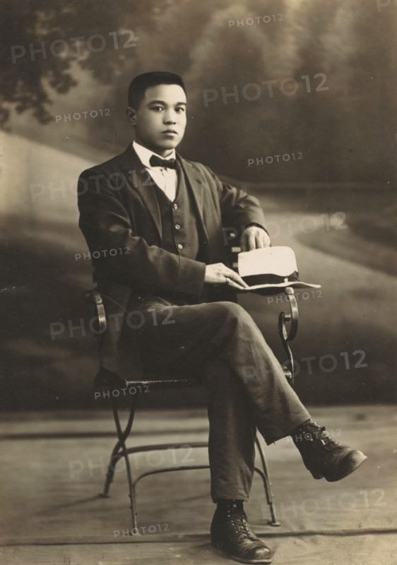 Portrait of an unknown Asian man, photograph, address: '????????????, ??????????? No 37, ???????? No 544', inscription: 'Huronán, Ban-uiu-ho ?', photographer: Chvunchva, Vladivostok, c. 1910 - c. 1930, Tsjvoenkhva, paper, pasteboard, baryta paper, gelatin silver print, developing gelatin silver print, height 144 mm x width 103 mm, height 170 mm x width 105 mm, adult man, anonymous historical person depicted, A seated young man in a dark three-piece suit and bow tie faces the camera with a neutral expression, one arm resting on the back of a metal chair and the other hand holding a light-colored hat placed on his knee, his legs crossed to show dark lace-up boots; the background is softly blurred with indistinct foliage and a path, and the image has a sepia-toned, vintage appearance.