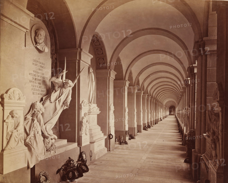 Genova Camposanto Galleria, Sculpture gallery with funeral wreaths at the Staglieno cemetery in Genoa, photograph, Part of French artist's album., Campo Santo (Genoa), c. 1880 - c. 1900, photographer, Genoa, paper, albumen print, height 218 mm x width 261 mm, gallery of sculptures, cemetery, park-like burying ground, A long covered corridor with repeating rounded arches and columns stretching into the distance, flanked by sculpted wall monuments and statues along one side, including a reclining angel figure and other carved human figures, a circular portrait relief high on the wall, decorative floral ceiling panels visible in some arch intrados, wreaths and floral arrangements placed at the bases of several monuments, a tiled or stone floor extending toward a small distant altar or niche, and a cloak-draped statue among the memorials.