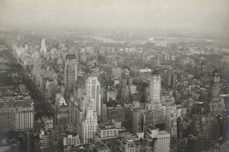 New York from RCA building uptown, New York seen from the RCA Building (now GE Building), part of the Rockefeller Center, United States, photograph, title: 'NEW YORK FROM, RCA BUILDING UPTOWN.', photographer: attributed to Wouter Cool, United States of America, 1936, North America, Attributed to, pasteboard, paper, gelatin silver print, developing gelatin silver print, height 157 mm x width 231 mm, height 315 mm x width 286 mm, city-view, and landscape with man-made constructions, skyscraper, A wide aerial view of a dense cityscape in grayscale showing many tall buildings of varying architectural styles and heights clustered close together, with streets and rooftops visible between them; a long tree-lined avenue extends from the foreground toward the left side, bordered by rows of buildings; distant low-rise areas and a waterway with bridges span the upper part of the scene, fading into a hazy sky above.
