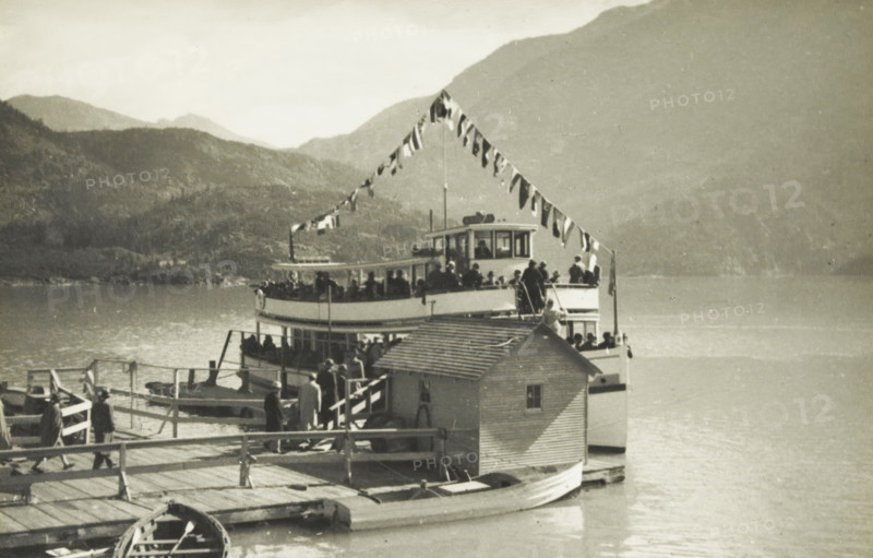 The Little Boat JD Ross, A boat on a lake at an unknown location in the United States, photograph, title: 'THE LITTLE BOAT, JD ROSS', photographer: attributed to Wouter Cool, United States of America, 1936, North America, Attributed to, pasteboard, paper, gelatin silver print, height 145 mm x width 220 mm, height 285 mm x width 315 mm, lake, traffic on the water, A dock extends into a calm body of water with a wooden walkway and railings leading to a larger dock structure where people stand and board a two-deck passenger boat decorated with a string of triangular flags draped from a mast to the bow. The lower deck of the boat is partially enclosed while the upper deck has railings and a small enclosed pilothouse with windows; silhouettes of people are visible on both decks and on the dock. A small shed with a slanted roof sits on the dock adjacent to the boat, and a rowboat is moored alongside the dock in the foreground. In the background, layered hills and mountains rise from the wate...