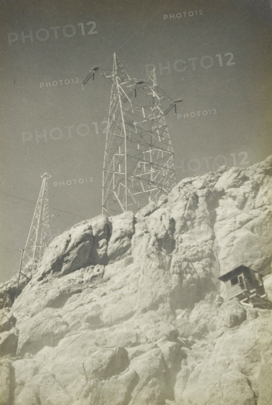 280 kv masts, Electricity pylons at an unknown location in the United States, photograph, title: '280 kV MASTEN', photographer: attributed to Wouter Cool, United States of America, 1936, North America, Attributed to, pasteboard, paper, gelatin silver print, developing gelatin silver print, height 228 mm x width 152 mm, height 315 mm x width 215 mm, pylons, power lines, A tall metal lattice tower with crossarms and insulators rises above a rocky cliff, alongside a smaller lattice tower visible further back, both set against a clear sky; the cliff shows rough, light-colored rock formations with cracks and shadows, and a small wooden structure is perched on the lower right face of the rocks.