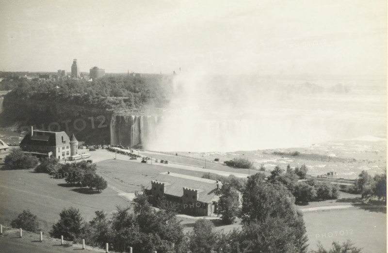 Niagara Falls (Horse Shoe), United States, Canada, Niagara Falls Horse shoe, photograph, title: 'NIAGARA FALLS, HORSE SHOE.', photographer: attributed to Wouter Cool, United States of America, 1936, North America, Attributed to, pasteboard, paper, gelatin silver print, developing gelatin silver print, height 154 mm x width 225 mm, height 285 mm x width 315 mm, waterfall, A wide aerial view shows a large waterfall with a heavy mist rising and spreading across the scene, cliffs and cascading water visible where the falls meet the river below. In the foreground there are grassy lawns dotted with trees, a roadway lined with parked cars and a few buses, and a substantial stone building with a steep roof and a small round turret near its corner. Nearby are smaller castle-like stone structures with battlemented tops, and a low stone wall edging the foreground. Beyond the falls a tree-covered ridge and a distant city skyline with several tall buildings and towers are visible under a pale sky.