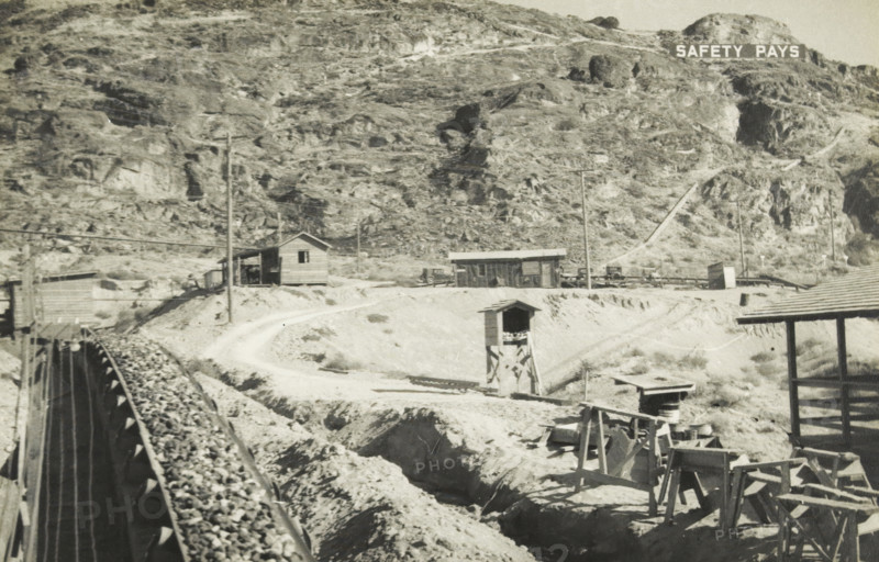Grand Coulee dam gravel supply to the concrete mixer, Grand Coulee Dam, Columbia River, United States: gravel supply, photograph, title: 'GRAND COULEE DAM, GRAVEL SUPPLY TO THE CONCRETE MILL', photographer: attributed to Wouter Cool, United States of America, 1936, North America, Attributed to, pasteboard, paper, gelatin silver print, developing gelatin silver print, height 150 mm x width 229 mm, height 315 mm x width 330 mm, dam (in canal), A sepia-toned landscape showing a rocky hillside with sparse vegetation and exposed rock faces, a narrow dirt road winding along the midground, small wooden buildings and sheds scattered near the road, utility poles and power lines crossing the scene, a raised wooden trestle carrying a conveyor or chute filled with rocks extending from the foreground toward the left, an open wooden tower structure standing near the road, assorted wooden scaffolding and crates in the lower right, and a hillside sign with block letters near the top edge.