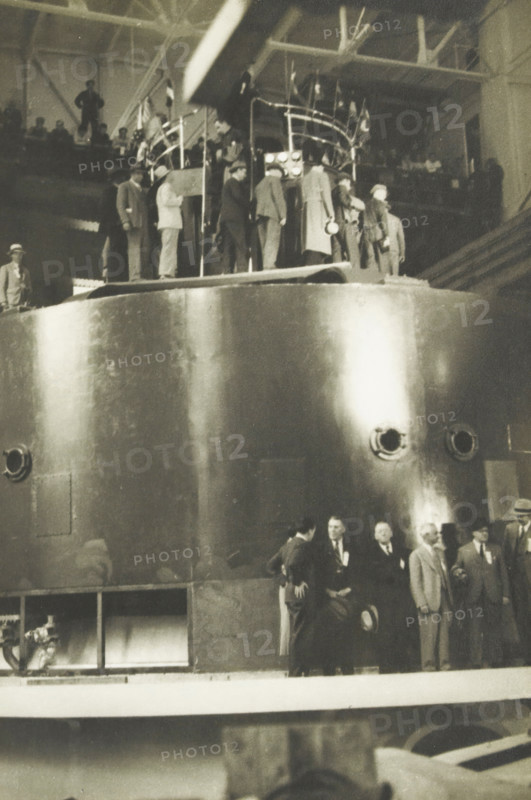 Water turbo of the Diablo Dam, Washington state, United States, Large water turbo 90, 000 kva central diabolo dam, photograph, title: 'LARGE WATER TURBORE 90, 000 kVA, CENTRAL DIABOLO DAM.', photographer: attributed to Wouter Cool, United States of America, 1936, North America, Attributed to, pasteboard, paper, gelatin silver print, developing gelatin silver print, height 227 mm x width 151 mm, height 315 mm x width 330 mm, dam (in canal), A large metallic cylindrical structure fills most of the image, its curved surface reflecting light and showing round openings and a small rectangular panel. Along the top edge of the cylinder a group of people stand on a platform, some wearing coats and hats and some clustered near a railing with vertical posts and lights. More people in suits and hats are visible at a lower level near the base of the cylinder, some standing and some seated, with a few facing each other in conversation. In the upper background an elevated walkway or balcony with str...