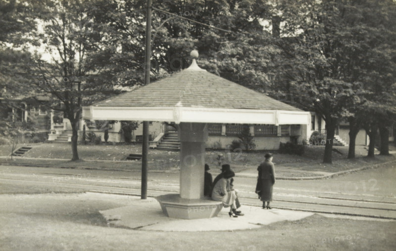 Tram stop in Seattle, Washington state, United States, Tram house in Seattle, photograph, title: 'TRAM HOUSE, IN SEATTLE', photographer: attributed to Wouter Cool, United States of America, 1936, North America, Attributed to, pasteboard, paper, gelatin silver print, developing gelatin silver print, height 142 mm x width 223 mm, height 315 mm x width 285 mm, tram, A grayscale photograph shows a sheltered street-side structure with a broad, shingled, pyramidal roof topped by a small finial, supported by a central column with a bench-like base. Several people are near and under the shelter; one person stands facing away from the camera while others sit on the bench, wearing long coats and hats. Behind the shelter is a tree-lined residential street with houses, porches, steps, and large trees with dense foliage. A lamppost stands next to the shelter and the foreground includes a paved area and streetcar tracks running parallel to the curb.