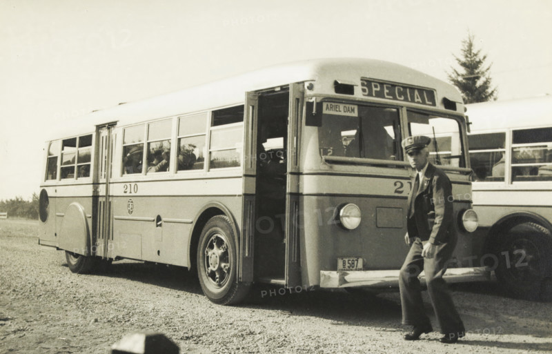 The newest buses, pneumatic brakes and gears, rear engine, Ariel Dam, Washington state, United States: bus, photograph, title: 'THE NEWEST BUSES, PNEUMATIC, BRAKES AND GEARS, REAR ENGINE.', photographer: attributed to Wouter Cool, United States of America, 1936, North America, Attributed to, pasteboard, paper, gelatin silver print, developing gelatin silver print, height 154 mm x width 228 mm, height 315 mm x width 285 mm, coach, A vintage bus parked on a gravel surface with its front door open and a man in a uniformed jacket and cap standing near the front bumper, the bus showing a row of windows along its side with passengers visible through them, round headlights on the front, large wheels with visible hubcaps, another similar bus partially visible behind it, and a tree in the background under a clear sky.