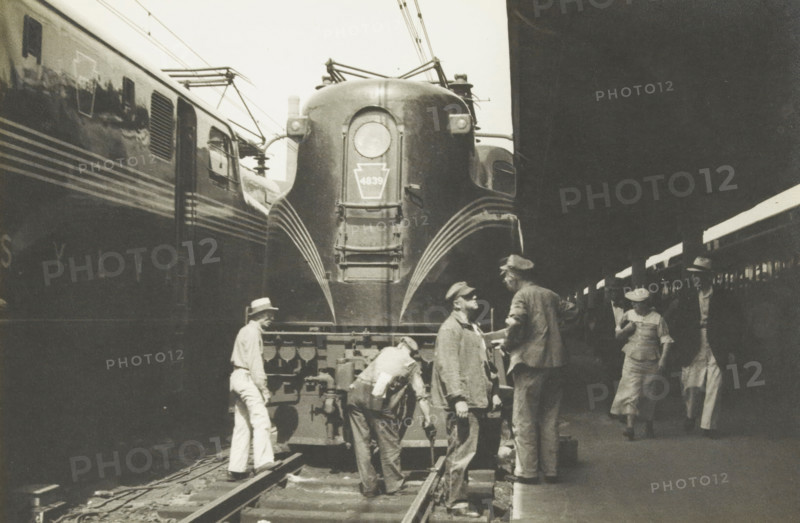 Electric locomotive of the Pensijlvania railroad, Electric locomotive of the Pennsylvania Rail Road, United States, photograph, title: 'ELECTRIC LOCOMOTIVE, OF THE PENSIJLVANIA RAILROAD.', photographer: attributed to Wouter Cool, United States of America, 1936, North America, Attributed to, pasteboard, paper, gelatin silver print, developing gelatin silver print, height 148 mm x width 220 mm, height 315 mm x width 285 mm, locomotive, A large streamlined train locomotive fills the center of the scene with curved metal surfaces and a prominent round headlight, flanked by parallel train cars on either side and overhead electrical wires. Several men stand and work on and around the front of the locomotive, wearing hats and work clothes, one leaning over near the coupling and another holding a tool while others converse. On the platform to the right a woman in a dress and hat walks while another man in a hat and long coat stands nearby carrying a bag. The image is monochrome and shows the p...