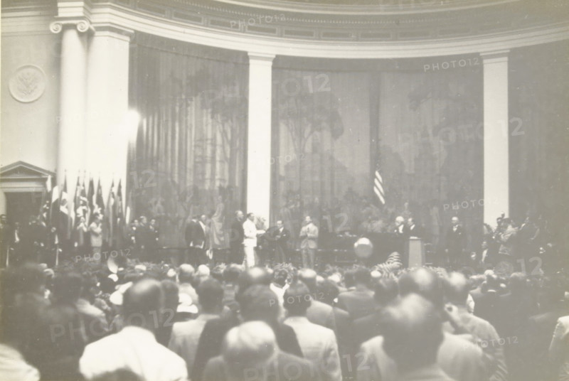 President Roosevelt enters the convention hall, Constitution Hall Washington, Speech by US President Franklin D. Roosevelt during the Third World Power Conference, Constitution Hall, Washington DC, United States, photograph, title: 'PRESIDENT ROOSEVELT ENTERS THE CONSCIENCE HALL, CONSTITUTION HALL WASHINGTON', photographer: attributed to Wouter Cool, United States of America, 1936-09-11, North America, Attributed to, pasteboard, paper, gelatin silver print, gelatin silver developing print, height 153 mm x width 228 mm, height 315 mm x width 285 mm, president, head of republic, A wide indoor scene showing a large audience in the foreground facing a raised stage area where a group of people stand and interact; tall columns frame a backdrop with a curtain and faint decorative patterns, and a display of vertical flags stands to one side while a single flag hangs near the center; several men in suits and one person in a light-colored outfit are positioned across the stage, with a podium and...