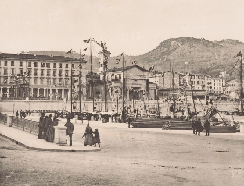 Le port, View of the harbor of Nice, page, photomechanical print, inscription: '35 Boulevard des Capucines Paris', inscription: 'Nice', photographer: Otto van Bosch, Nice, photographer: Paul Boyer (1861-1908), cliché maker: Dujardin, printer (person): Chardon-Wittmann, c. 1888 - in or before 1890, printer (person), Provence-Alpes-Côte d'Azur, paper, photoaquatint, height 186 mm x width 233 mm, Print after page 135., harbour, A waterfront scene showing a promenade with a stone balustrade and lampposts, people in period clothing walking and standing in groups, and a cluster of masts and rigging from moored sailing vessels decorated with flags and pennants. Behind the boats a long multi-story building with many windows and balconies faces the water, alongside a clock tower and additional smaller stone buildings. Stacked timber and cargo are visible near the quayside, and hills covered in vegetation rise in the background. The image has a monochrome, sepia-toned appearance.