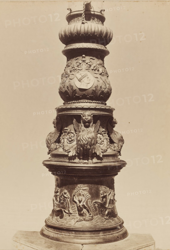 Relief-decorated column, photograph, photographer: attributed to Carlo Naya, c. 1880 - c. 1900, Attributed to, pasteboard, paper, albumen print, height 358 mm x width 272 mm, height 650 mm x width 500 mm, A tall ornate column segment with a rounded top section featuring fluted and beaded molding, a central bulbous urn-like form decorated with floral motifs and a raised circular medallion showing a profile of a human head, and a lower band with sculpted winged lion figures and elaborate foliage; beneath that a cylindrical base carved with relief scenes of human figures in dynamic poses among waves and leafy ornamentation, all rendered in a uniform stone or metal material against a plain light background.