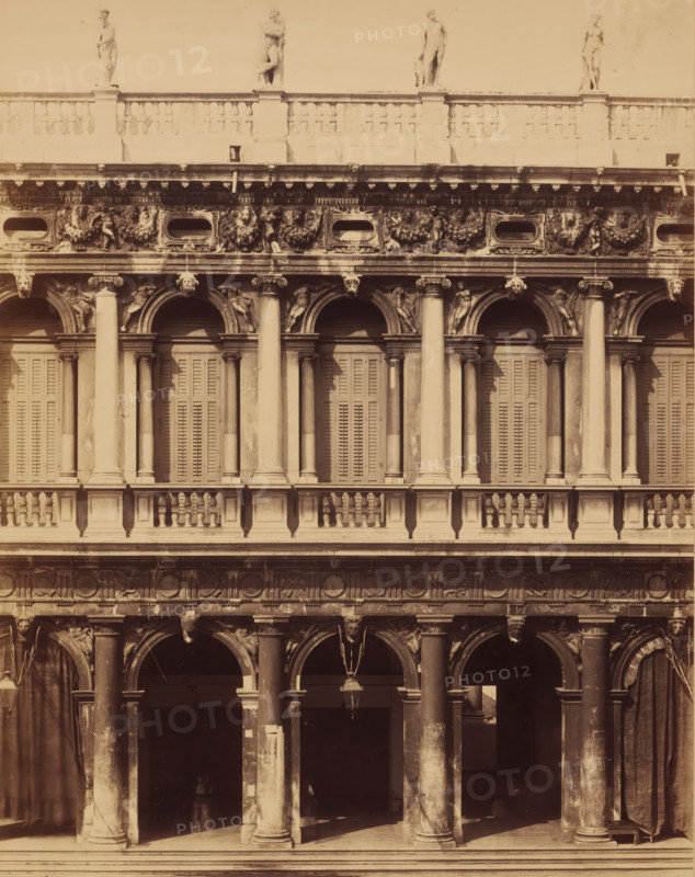 Palace opposite the Doge's Palace in Venice, photograph, photographer: attributed to Carlo Naya, c. 1850 - c. 1900, Carlo Naya, photographer, Attributed to, paper, pasteboard, albumen print, height 352 mm x width 270 mm, height 650 mm x width 270 mm, Venice, A multi-level stone façade with arched openings and tall columns, decorative capitals and balustrades, shuttered windows behind the arches on the upper level, sculpted reliefs and garlands in the horizontal frieze, carved human figures and heads integrated into the ornamentation, statues standing along the roofline, a hanging lantern beneath one of the arches, heavy curtains or drapery at the sides of the lower arches, weathered surfaces and patches of darker staining on the stone.