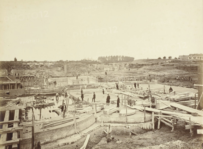 Construction of Locks at Veere. Possibly No. 10 Lock chambers and inner heads of the double Lock at Veere, viewed from the pier between the outer heads, Veere, lock under construction, photograph, photographer: Pieter Oosterhuis, 1871-08, pasteboard, paper, albumen print, height 320 mm x width 419 mm, weigh-house, A wide construction site with exposed foundations, wooden planks, beams, and scaffolding stretching across the foreground and middle distance, with workers scattered on platforms and among the construction materials; shallow water pooled in trenches and channels near the front, piles of stone and rubble, and various tools and carts visible; earthworks and mounds of soil rise toward the background where stacks of logs and stone blocks sit behind a low ridge; a small grove of trees and a few houses with pitched roofs appear on the horizon; the sky above is pale and mostly empty.