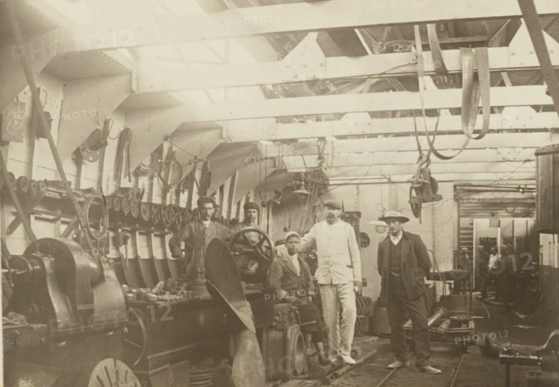 Machine shop, photograph, Interior of a machine shop. Photo pasted on cardboard, without texts. Part of the folder with 44 photos of dredgers from the company A. Bos & JP Heijblom in Dordrecht., c. 1900 - c. 1910, photographer, paper, pasteboard, gelatin silver print, developing gelatin silver print pasted on cardboard, height 122 mm x width 171 mm, height 240 mm x width 298 mm, Person, Firma A. Bos & JP Heijblom, factory interior, dredge, dredging machine, A sepia-toned interior shows a workshop with overhead beams and hanging straps and pulleys, a row of mounted wheels and machinery along the left wall, large gears and a propeller blade in the foreground, workbenches with tools and parts, and five men standing and seated near the machines wearing work clothes, a suit with coat and hat, and a light-colored uniform; the ceiling has structural supports and lighting, and the background contains more equipment and a doorway leading to another room.