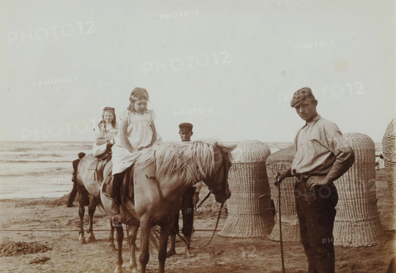 At the Beach, photograph, inscription: 'H + Nan, zeev, ...', photographer: attributed to Willem de Jong, Netherlands, photographer: attributed to Piet de Jong, c. 1900, Attributed to, paper, baryta paper, daylight collodion silver print, height 90 mm x width 120 mm, Person, Johanna Elisabeth Borgerhoff Mulder-Vreede (Nan), Johanna Petronella Lambertina Stokhuyzen-de Jong (Hans), beach, riding a horse, ass, or mule, rider, horseman, child, North Sea, A sepia-toned beach scene shows two young girls sitting on horses facing forward, each wearing light dresses and patterned headscarves, with one girl nearer the camera looking down and the other slightly behind smiling. A man stands to the right wearing a cap, a long-sleeved shirt with sleeves rolled, striped trousers, and holding a walking stick while looking toward the camera. Another man in a dark cap stands between the horses, partially obscured. The horses have light manes and saddles, and large woven wicker baskets or beach chairs sta...