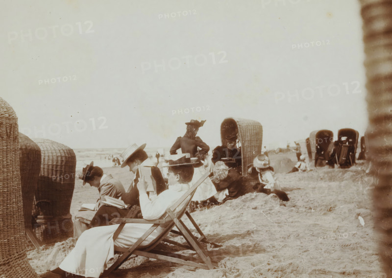 At the Beach, photograph, photographer: attributed to Willem de Jong, Netherlands, photographer: attributed to Piet de Jong, c. 1895 - c. 1920, Attributed to, paper, baryta paper, daylight collodion silver print, height 89 mm x width 120 mm, open-air recreation, beach, reading, North Sea, A sepia-toned beach scene showing people seated on folding deck chairs and on the sand, many wearing wide-brimmed hats and Victorian-style clothing, with woven wicker beach shelters arranged along the shore and one wicker shelter opened facing the camera, groups of adults and children clustered together, a person in a dark jacket and hat standing with hands on hips near the center, and a textured foreground of sand and low dunes.