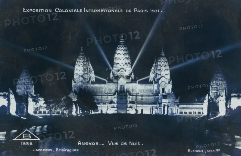 Exposition Coloniale Internationale de Paris 1931. Angkor - Vue de Nuit, View of the reconstructed temple city of Angkor at the Colonial Exhibition of Paris in 1931, at night, photograph, picture postcard, negative number: 'B 1236', Paris, publisher: Adolphe Braun & Cie., France, c. 1931-05 - c. 1931-11, photographer, paper, gelatin silver print, developing gelatin silver print, height 90 mm x width 137 mm, world fair, world exhibition, World Exhibition 1931 (Paris), A nighttime scene of an illuminated temple complex with a central stepped entrance flanked by ornate towers and smaller structures, bright spotlights casting beams upward and outward from the temple, reflective water or dark foreground in front of the complex, silhouetted trees and sculptural forms at the base of the temple, and a dark sky above.