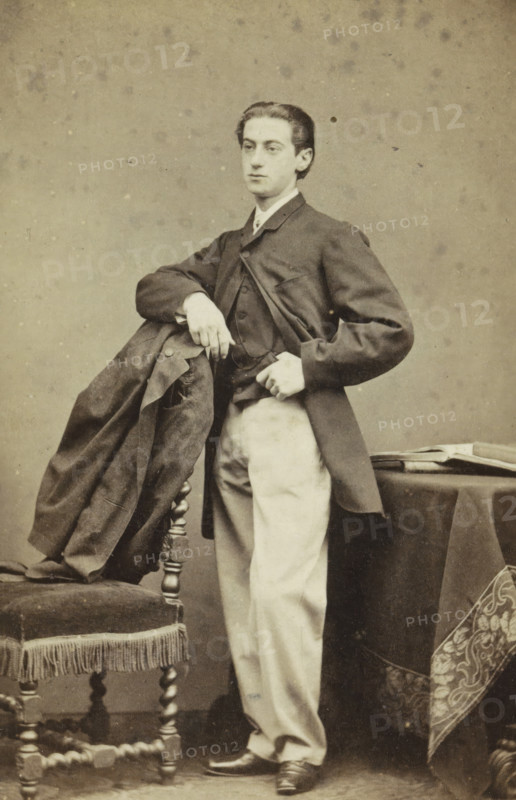 Portrait of an Unknown Young Man, carte-de-visite, name: 'Wegner & Mottu photo. Amsterdam', photographer: Wegner & Mottu, Amsterdam, c. 1865, height 89 mm x width 57 mm, height 105 mm x width 62 mm, historical persons not known by name, A sepia-toned portrait of a young man standing with one elbow resting on the back of an upholstered chair draped with a dark coat, wearing a dark jacket over a lighter waistcoat and light-colored trousers, his hair neatly combed back and his expression neutral, one hand resting on his hip and the other holding the coat, a small table covered with a dark cloth and a folded cloth with a patterned border visible beside him, and the background showing a plain mottled backdrop.
