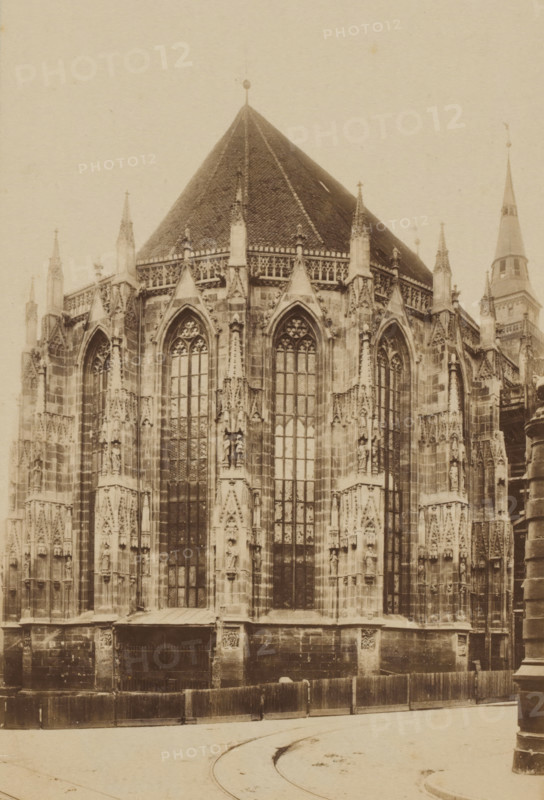 St. Sebald Nürnberg, photograph, c. 1850 - c. 1900, photographer, paper, albumen print, albumendruk, height 162 mm x width 104 mm, height 182 mm x width 134 mm, Bavaria, Nuremberg, St. Sebaldus Church, A sepia-toned photograph of a large Gothic church exterior showing a pointed tiled roof and tall ornate windows with pointed arches, tracery, and mullions; the stone facade features vertical buttresses, pinnacles, carved niches with statues, decorative stone carvings, and sculpted details; a rounded spire is visible to one side, and temporary wooden fencing runs along the base of the building in front of a stone plinth; the foreground shows a curved roadway with faint tram tracks and a cylindrical street fixture at the near right edge.