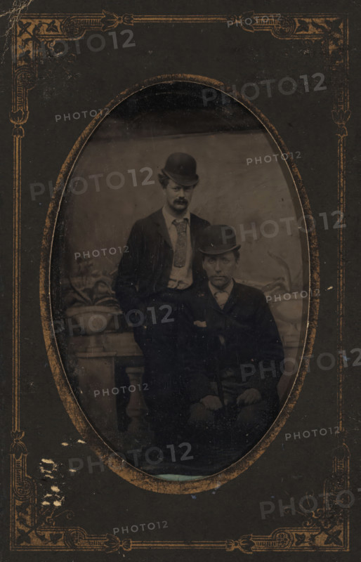 Portrait of Willem Kloos and Hein Boeken, photograph, photographer: unknown, Netherlands, c. 1885 - c. 1890, metal, inorganic material, iron (metal), paper, ferrotyping, ferrotype in paper cover, height 81 mm x width 53 mm, height 118 mm x width 78 mm, Person, Willem Kloos, Hein Boeken, historical persons, head-gear: hat, An oval-framed black-and-white photograph set within a dark decorative mat shows two men wearing dark suits and bowler hats; one man stands behind the other who is seated, both with neutral expressions, the standing man has a patterned necktie and light-colored shirt visible beneath his jacket while the seated man wears a darker necktie and holds a cane across his lap, a potted plant on a pedestal appears beside them and a plain backdrop fills the space behind, the mat around the oval has worn gold ornamental corner designs and scuffed edges.