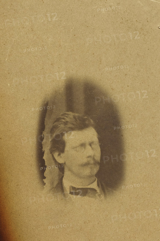 Portrait of Theodorus Kroes, photograph, number: '95', photographer: attributed to Antoon Bauduin, 1862 - 1866, Attributed to, paper, albumen print, albumendruk, height 99 mm x width 72 mm, historical persons (portraits and scenes from the life), A faded sepia-toned photograph showing the upper portion of a man's head and shoulders emerging from a darker oval vignette against a lighter background, with the man wearing a dark bow tie and a high-collared shirt, short wavy hair, and a mustache; the left edge of the image has a torn, darkened area with irregular burn-like edges and a rough white torn corner.