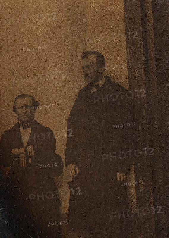 Self-portrait with unknown man (left), Two white men, one sitting one standing (former title), photograph, number: '73', photographer: attributed to Antoon Bauduin, 1862 - 1866, Attributed to, paper, albumen print, height 135 mm x width 92 mm, historical persons not known by name, A sepia-toned photograph shows two men standing against a plain backdrop, one shorter man seated or positioned slightly lower on the left wearing a dark suit with a bow tie and holding an object at chest height, and a taller man on the right wearing a dark coat with hands resting near his sides; the image has dark irregular burn-like edges and visible creases and discoloration across the surface.
