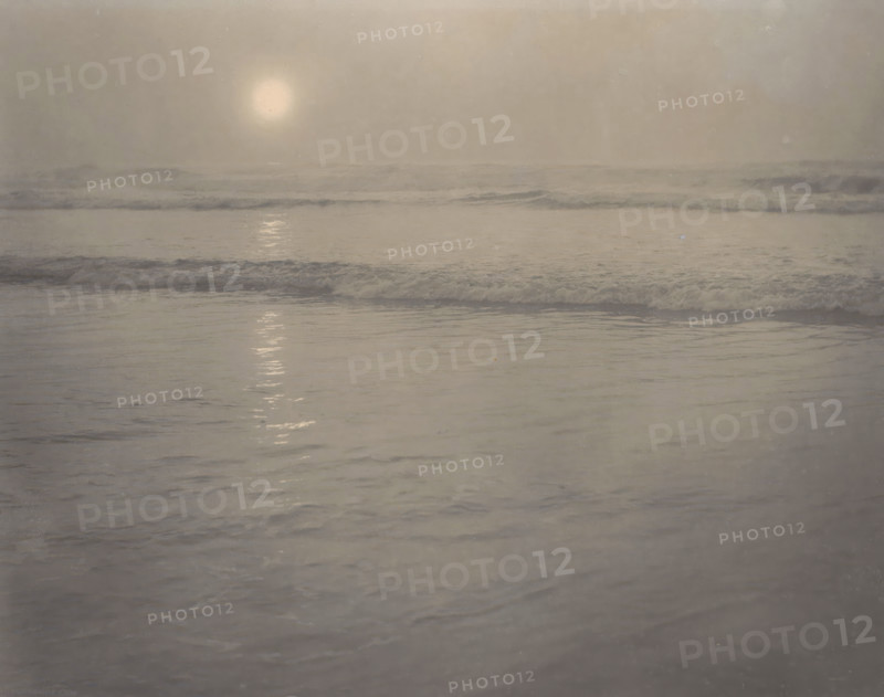 Seascape, photograph, photographer: Rudolf Eickemeyer, 1900 - 1906, pasteboard, paper, platinum process, height 184 mm x width 234 mm, height 278 mm x width 432 mm, ocean, A muted seascape with a pale disc of light low in a hazy sky reflecting as a vertical shimmer on calm water, gentle wavelets and foamy surf stretching across the midground, and smooth rippled water in the foreground, all rendered in soft gray and beige tones.