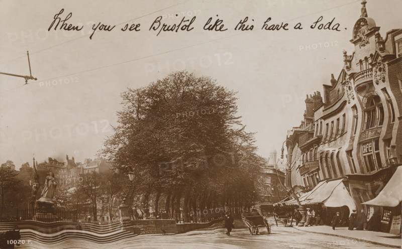 When you see Bristol like this have a Soda, Rotary 'Grotesque' Series, photograph, picture postcard, Bristol, publisher: Rotary Photographic Co., London, 1905 - 1920, photographer, paper, baryta paper, daylight gelatin silver print, height 85 mm x width 136 mm, drunkenness, street (city(-scape) with figures, staffage), A sepia-toned street scene shows a row of tall leafy trees along a sidewalk, a statue on a pedestal within a fenced area to the left, and ornate multi-story buildings with decorative facades and awnings lining the right side; people in period clothing stand and walk along the street, a handcart is visible near the center, street lamps and railings appear along the avenue, and overhead lines cross the pale sky.